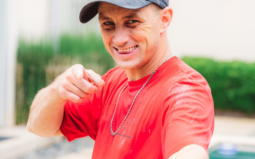 A Tulsa Interior Painter in a red shirt splattered in paint, wearing a blue hat, smiles and points at the camera.