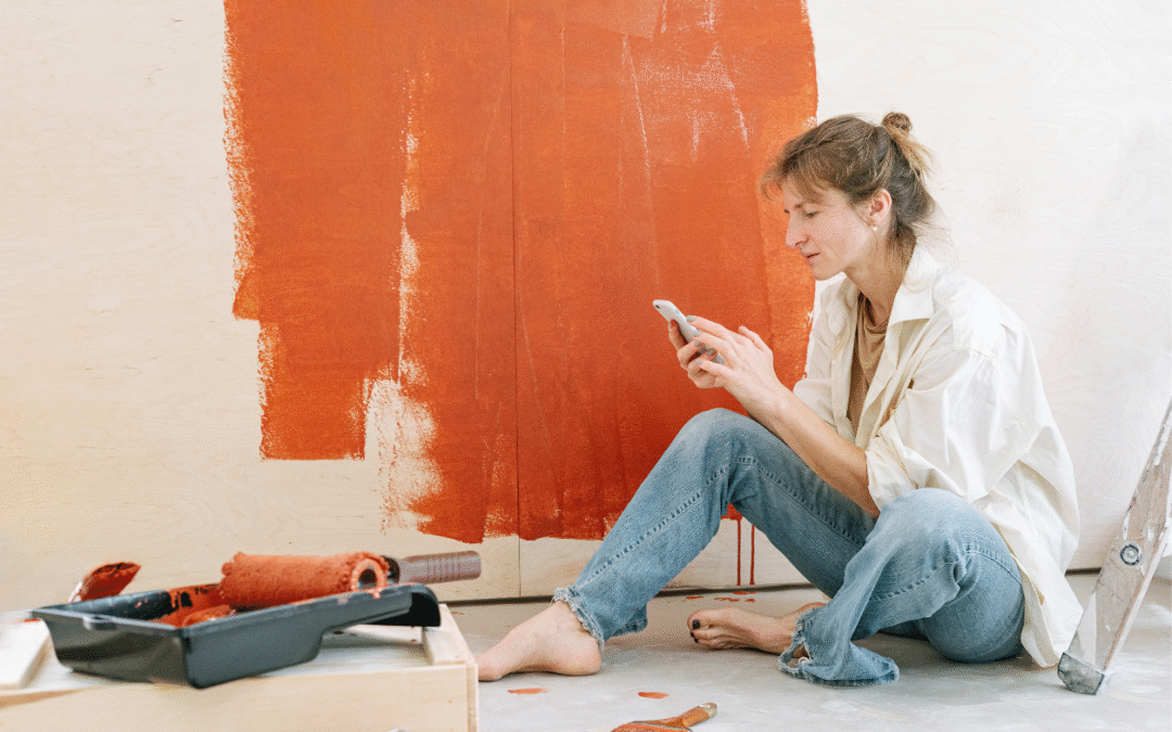 Woman sitting on the ground on her phone taking a break from painting the wall