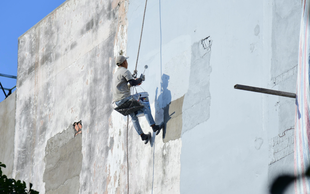 Man painting a cement wall in a harness with ropes attached