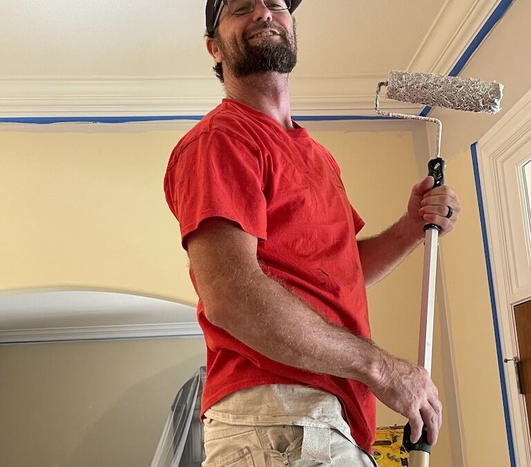 An Interior Painter Tulsa in a red shirt smiles at the camera with a roller in his hand.