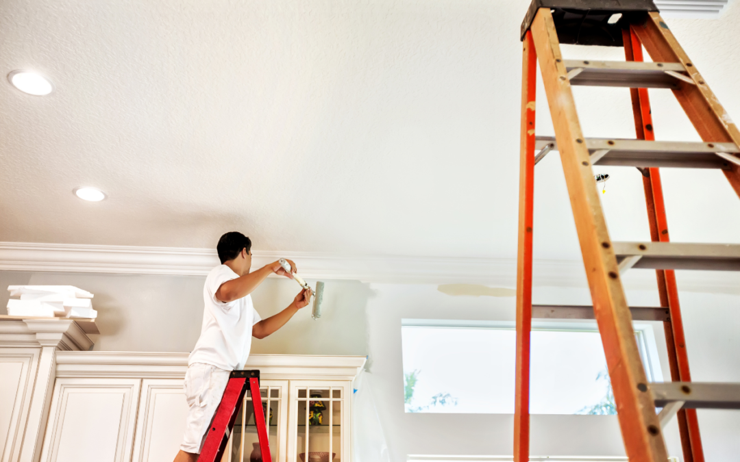 A person wearing white work clothes is standing on a red step ladder, painting the upper wall near the ceiling inside a bright, modern room. They’re using a paint roller to apply a light-colored paint along the wall just below the ceiling trim. The room features recessed ceiling lights, white cabinetry, and large windows letting in natural light. A tall extension ladder stands upright on the right side of the image, suggesting an ongoing professional painting job or home renovation.